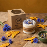 Beauty Scents Large Candle With Wild Flowers next to its branded box surrounded with corn flowers on a wooden table.