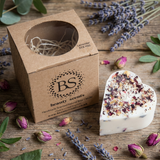 Beauty Scents Small Heart Candle With Rose Petals next to its branded box on the wooden table surrounded by dried herbs