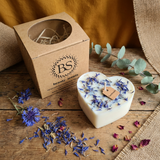 Heart-shaped candle with dried flowers on a wooden surface next to a cardboard box labeled 'BS beauty scents'.