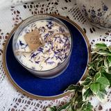 Large glass candle topped with dried wildflowers and a rustic tag, displayed on a dark blue plate with greenery and lace background.