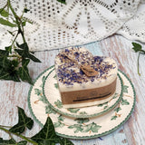 Large heart-shaped wild flower candle with kraft label, decorated with dried blue petals, on vintage ivy plates and rustic wood background