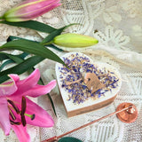Large heart-shaped wild flower candle with kraft label, decorated with dried blue petals, next to pink lilies on lace tablecloth