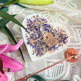 Large heart-shaped wild flower candle with kraft label, decorated with dried blue petals, next to pink lilies on lace tablecloth