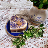 Large heart-shaped wild flower candle with kraft label, decorated with dried blue petals, on vintage ivy plates and rustic wood background