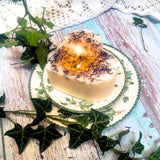 Large heart-shaped wild flower candle with kraft label, decorated with dried blue petals, on vintage ivy plates and rustic wood background