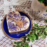 Heart-shaped candle with dried flowers on a blue plate, surrounded by green leaves and a lace tablecloth.