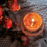 Medium rose petal candle burning on vintage plate, surrounded by pink roses and lace tablecloth in warm evening light