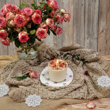 Candle with rose petals on a lace tablecloth next to a vase of pink roses against a wooden background