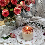 Medium rose petal candle burning on vintage plate, surrounded by pink roses and lace tablecloth in warm evening light
