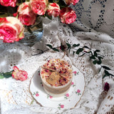 Medium candle with rose petalswith floral elements on a decorative plate, surrounded by pink roses and green leaves on a lace tablecloth.