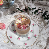Small glass rose petal candle burning on vintage floral plate, surrounded by pink roses and lace tablecloth