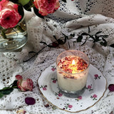 Candle in a glass holder with rose petals on a decorative plate, surrounded by flowers and lace fabric.