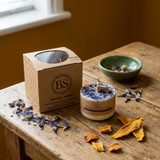 Candle with 'BS' branding on a wooden surface with dried flowers and a box.