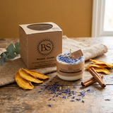 Beauty Scents candle with packaging on a wooden surface with dried leaves and cinnamon sticks.