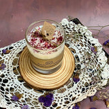 Small glass rose petal candle with kraft label on wooden slice, vintage lace and dried purple flowers, rustic mauve background