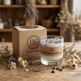 Handmade Beauty Scents glass candle decorated with wildflowers, displayed with its box and botanical elements on a wooden table in an artisan workshop.