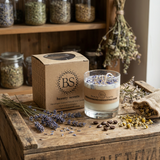 Large glass candle with wildflowers on a rustic wooden table, surrounded by dried herbs, lavender, and natural ingredients in a cozy workshop setting.