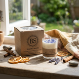 Small glass candle with blue wildflowers and a kraft box on a sunny windowsill, surrounded by dried orange slices, cinnamon sticks, and a garden view.