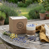 Small glass candle with blue wildflowers and a kraft box on a rustic garden table, surrounded by fresh chamomile, lavender, and a yellow cloth with greenery in the background.