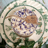 Small handmade wild flower candle with kraft label, decorated with dried blue petals, on vintage ivy plates and rustic wood background