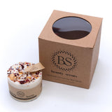 A small scented soy wax candle with rose petals on top, next to its brown cardboard box with the brand 'Beauty Scents' written on it.